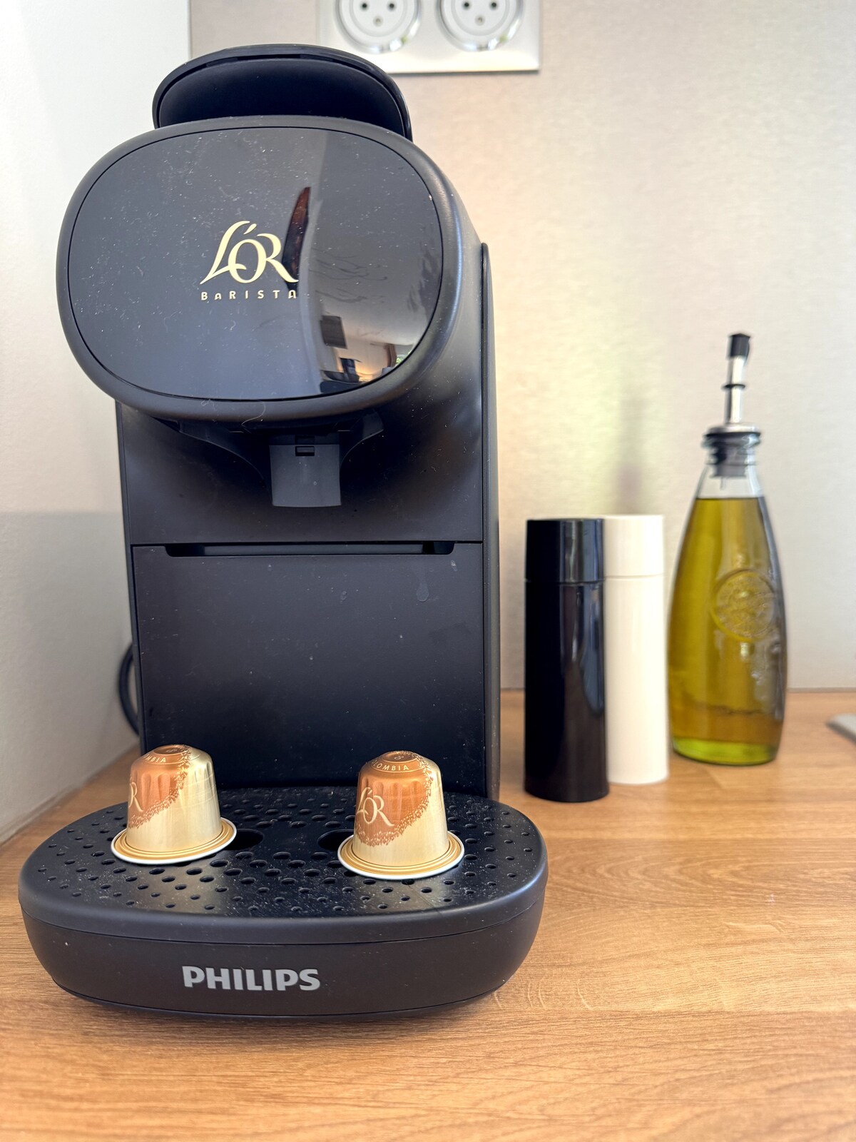 A sleek coffee machine is positioned on a wooden countertop, featuring two coffee capsules waiting to be used. Nearby, a black pepper grinder, a white container, and a bottle of olive oil are visible, emphasizing a well-equipped kitchen area.