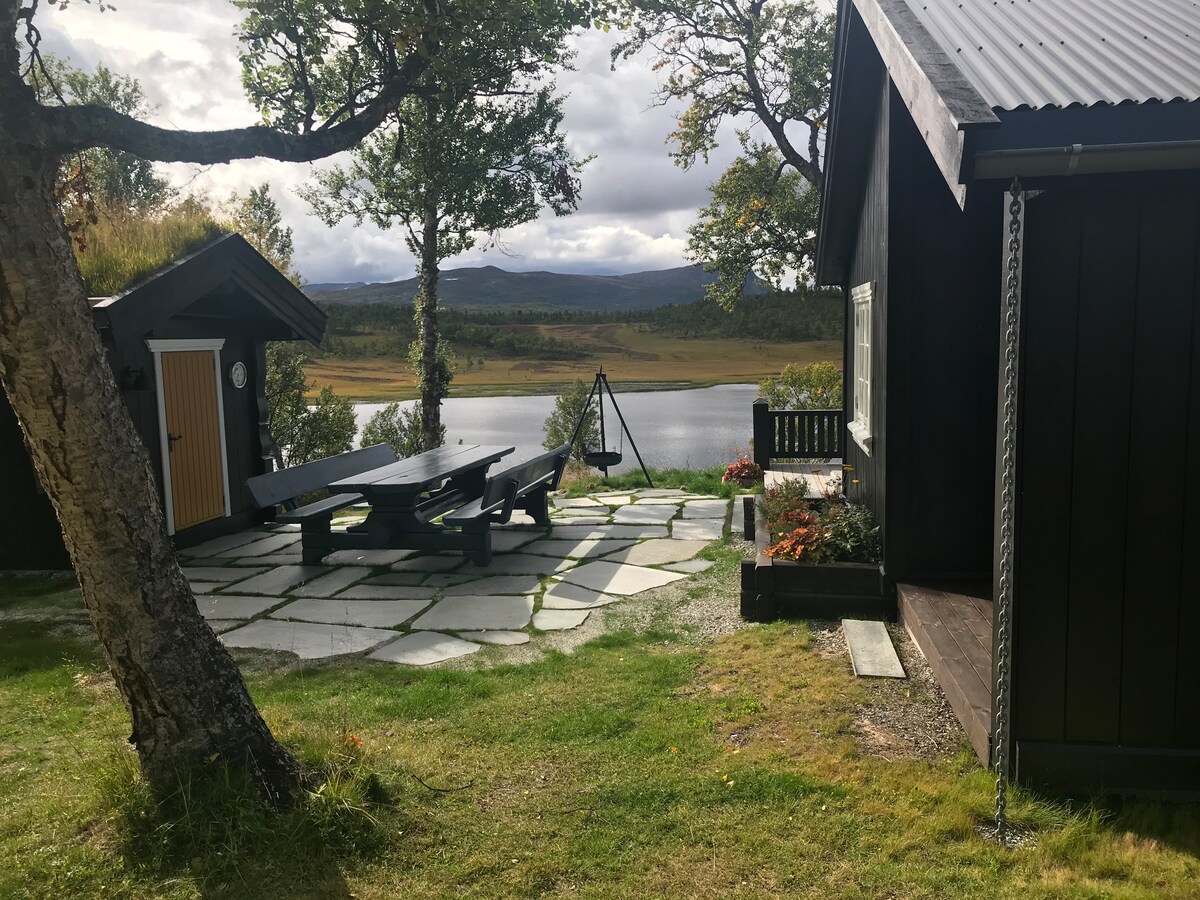 A stone patio area features a large table surrounded by benches, overlooking a tranquil lake. Two wooden structures in contrasting colors are visible, with trees and grassy areas framing the scene. The landscape beyond showcases rolling hills and a cloudy sky.