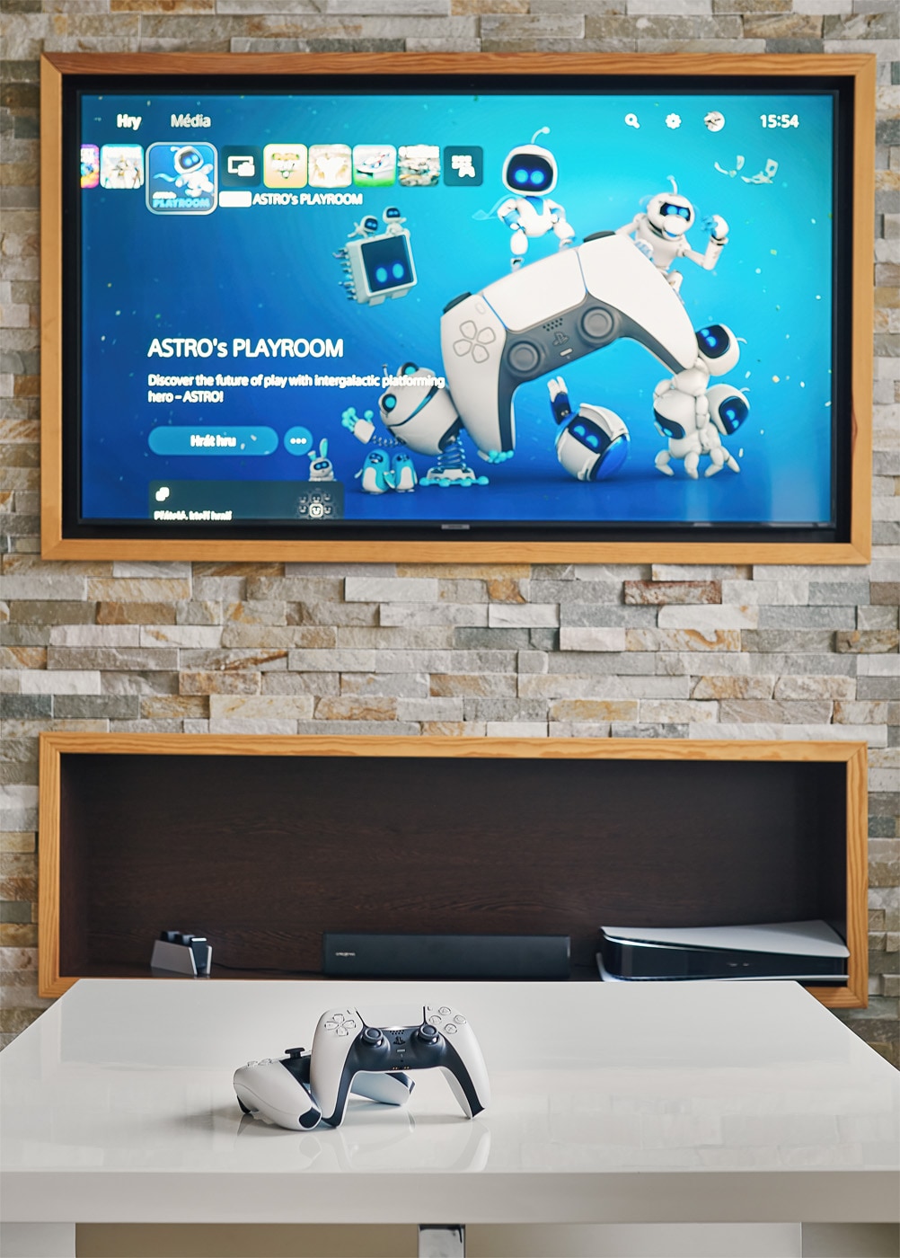 A flat-screen television mounted on a stone wall displays a gaming interface featuring colorful graphics. A modern entertainment unit below holds a video game console, while a game controller rests on a sleek white coffee table, inviting guests to engage in leisure activities.