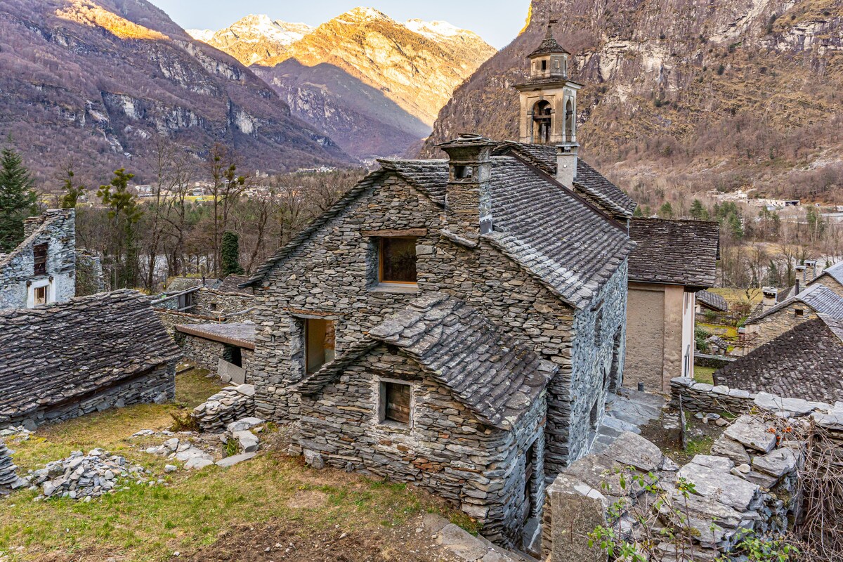 The exterior of the historical stone house is visible, showcasing its traditional architecture. The surrounding landscape features mountains in the background, while the house highlights a sloped roof and window details. A bell tower rises above, adding to the rustic charm of the structure.