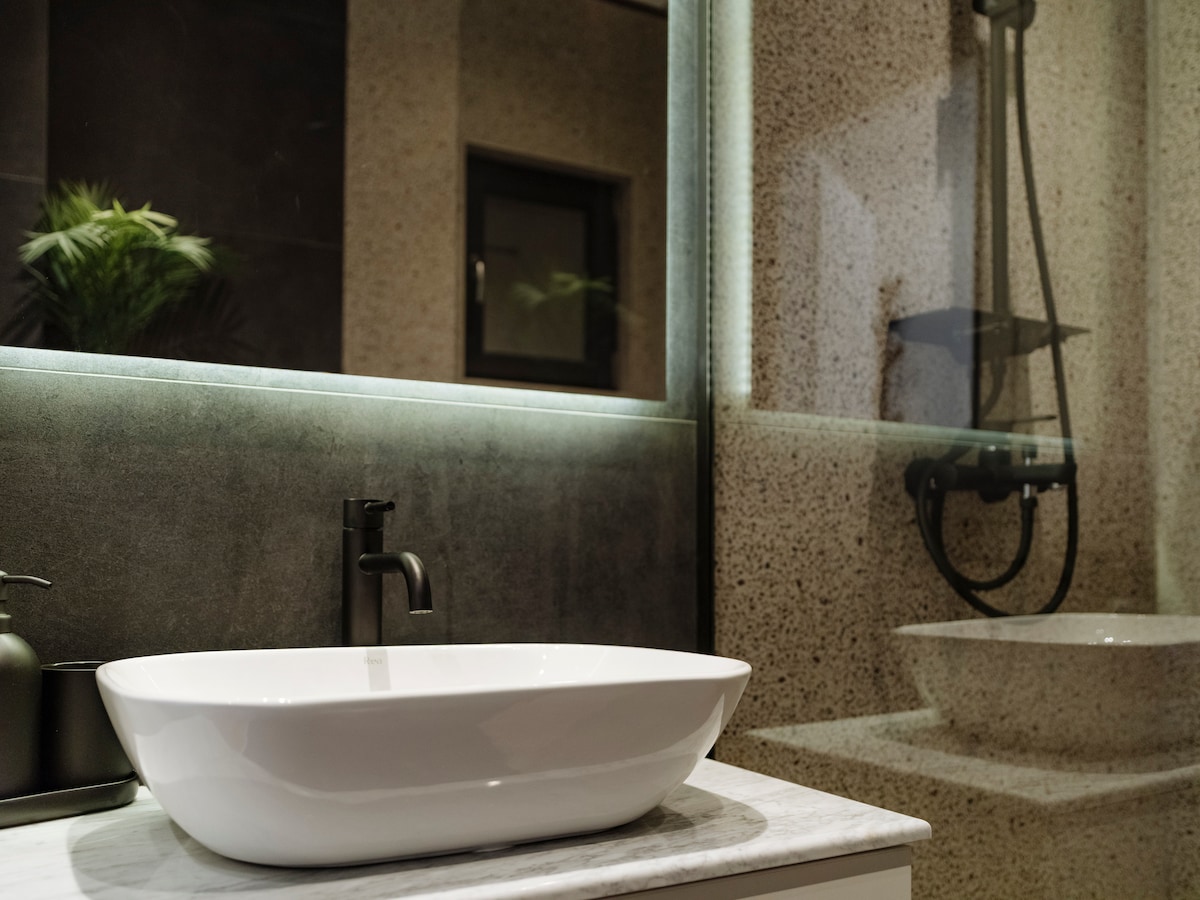 A modern bathroom features a sleek countertop with a spacious white basin and a minimalist faucet. The shower area is enclosed in glass, reflecting light from the well-lit mirrors. Subtle greenery is visible through a small window, contributing to a refreshing ambiance.