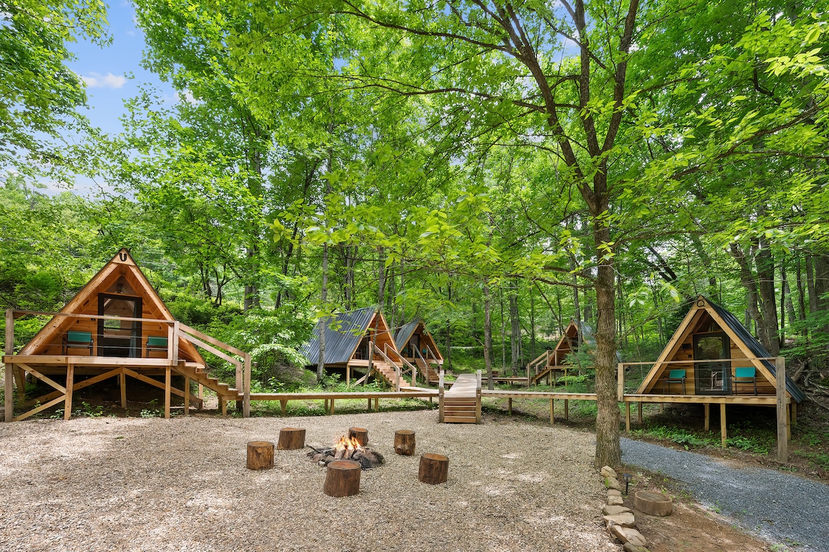 A cluster of A-frame cabins is nestled among lush green trees, featuring wooden decks and connected by boardwalks. A fire pit sits in the center of the area, surrounded by seating, inviting relaxation and community gatherings.