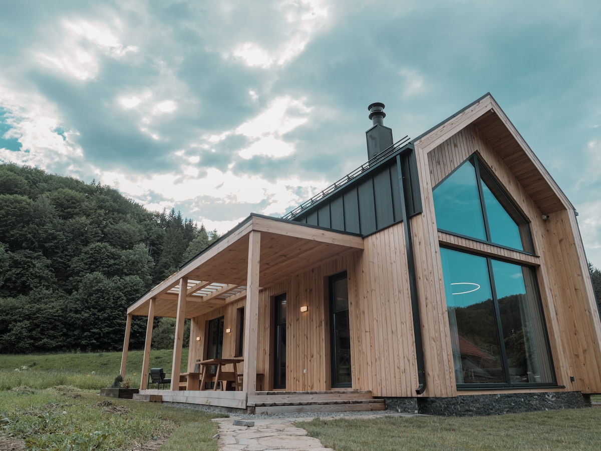A wooden structure with large glass windows reflects the nearby landscape. The peaked roof and expansive porch provide an inviting outdoor space. A stone pathway leads to the entrance, surrounded by greenery and under a cloudy sky.