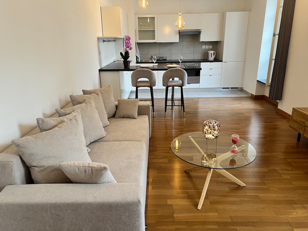 The living area showcases a light-colored sofa adorned with soft cushions. A glass coffee table with a decorative centerpiece is positioned in front. In the background, a modern kitchen features sleek cabinetry and a bar area with two stools, all set against polished wooden flooring.