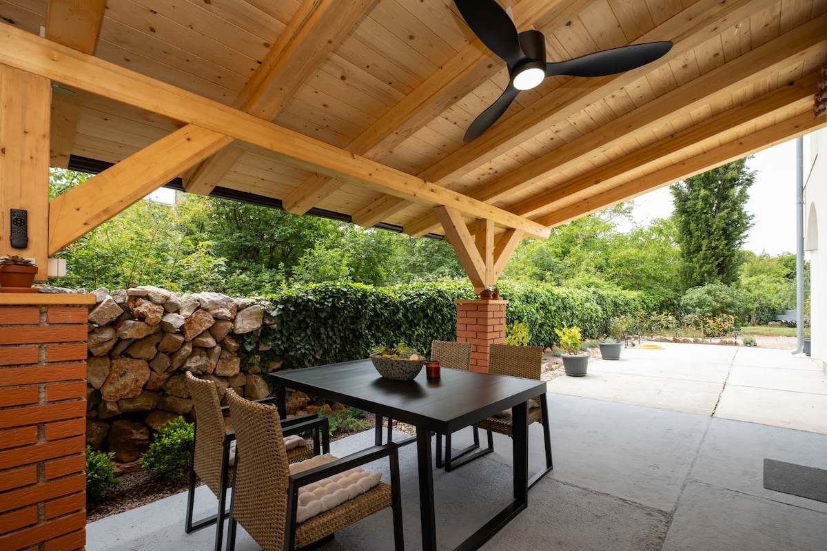 An outdoor covered patio is visible, featuring a dark table surrounded by four wicker chairs. A ceiling fan is mounted above, and greenery is seen in the background, adding to the serene outdoor space. Natural stone and lush plants frame the area.