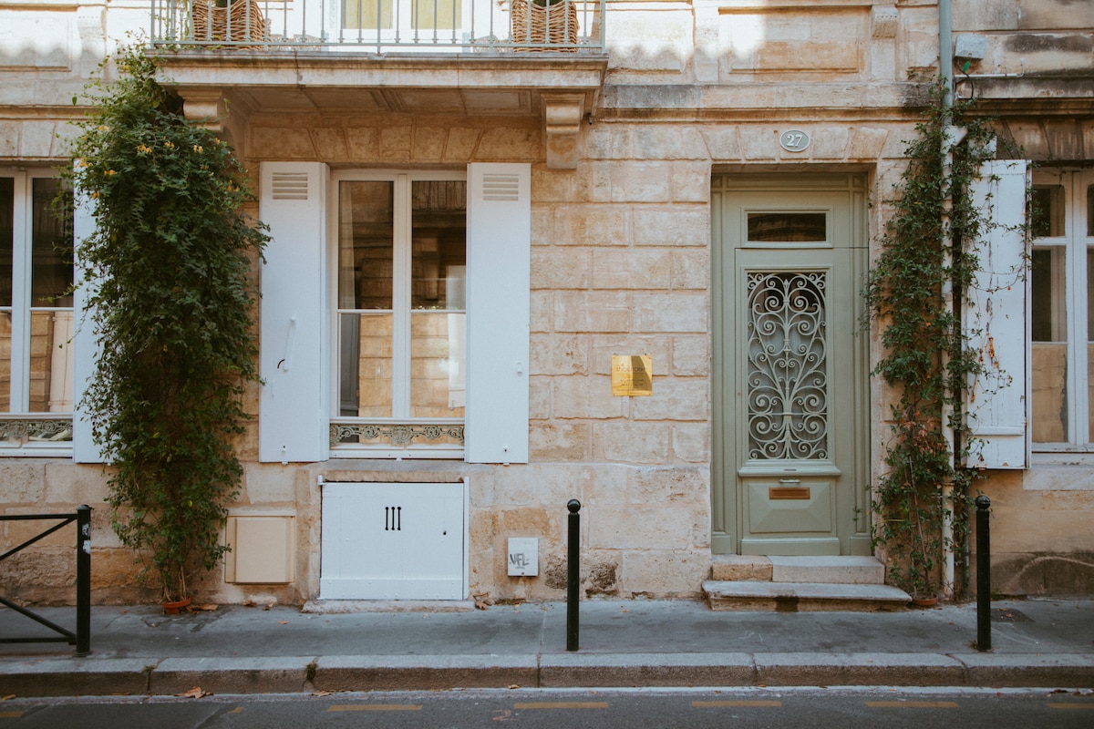 The exterior of a charming stone building features intricate ironwork surrounding the entrance door. Light-colored shutters flank the tall windows, with greenery climbing the wall. A textured facade enhances the traditional architecture, while a small stairway leads up to the inviting door.