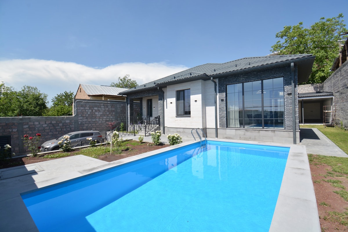 A modern single-story villa is shown, featuring large windows that allow natural light to illuminate the interior. A bright blue pool reflects the sky, surrounded by well-maintained landscaping. The driveway is visible, providing parking access beside the villa.