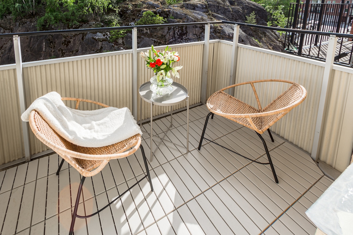 A private balcony is presented with two woven chairs and a small glass table. A vase filled with flowers adds a touch of color. Sunlight illuminates the space, highlighting the wooden flooring and the surrounding greenery.