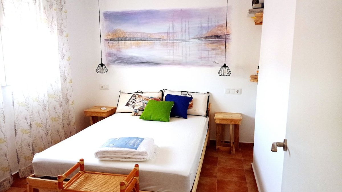 A bright bedroom features a spacious bed adorned with colorful decorative pillows. A large artistic mural of a serene landscape is displayed on the wall above. Two wooden nightstands flank the bed, while a small woven stool adds a natural touch. Soft light filters through the curtain-covered window.