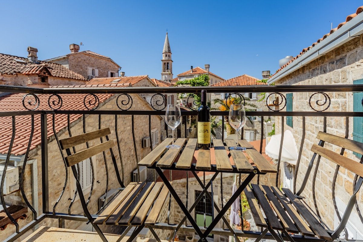 A small balcony features a wooden table with two chairs, accompanied by a bottle of wine and glasses. In the background, rooftops of old buildings and a church tower are visible under a clear blue sky.