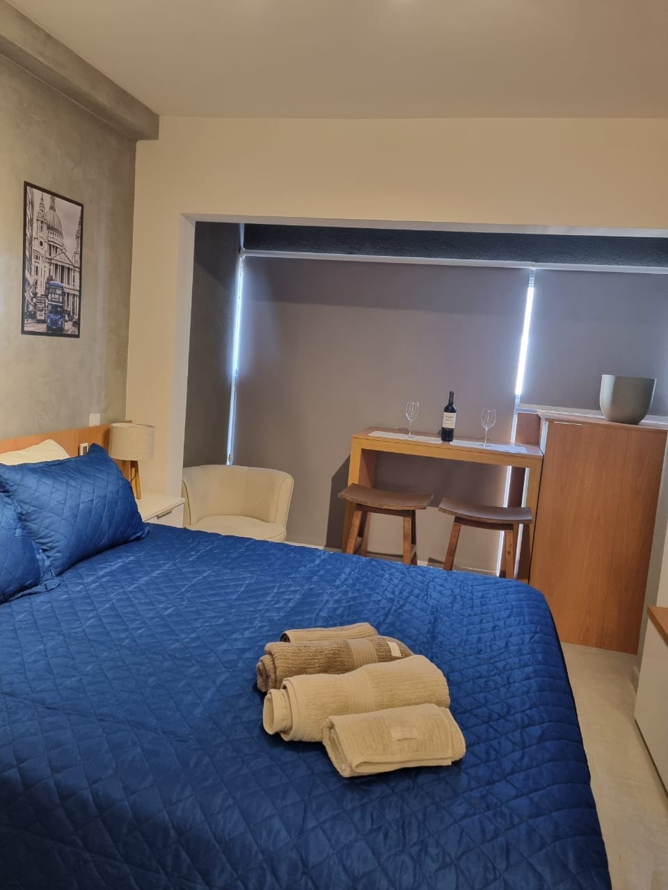 A spacious bed with a deep blue coverlet is complemented by neatly arranged towels in neutral colors. A small table set with two glasses and a wine bottle is visible, alongside a cozy armchair and partially illuminated by natural light from a nearby window.