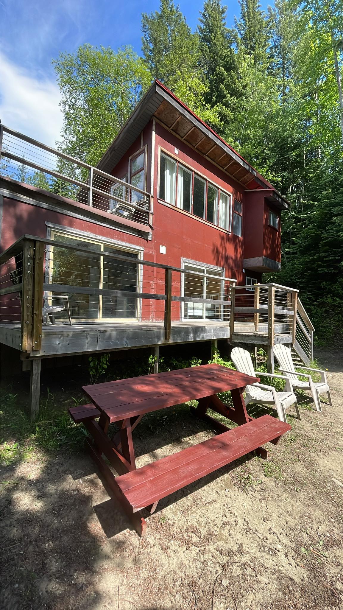 A red two-story cabin is framed by tall trees, featuring a spacious deck with glass railings. A picnic table is positioned on the ground level alongside several lawn chairs, inviting outdoor relaxation. Natural surroundings highlight the cabin's connection to the landscape.