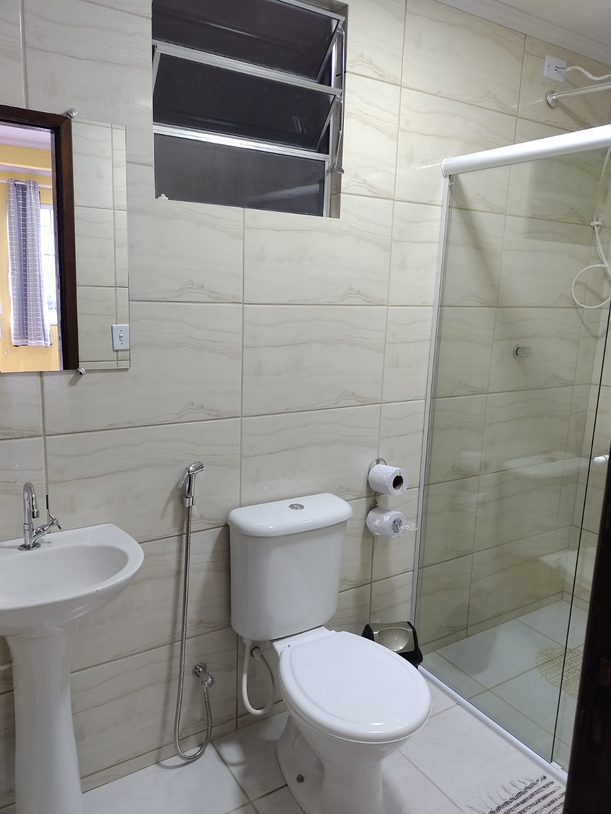 The bathroom features modern fixtures, including a white sink and toilet. A glass shower is present, allowing easy access. Neutral-toned tiles cover the walls, and natural light enters through a small window above the shower, enhancing the overall brightness of the space.