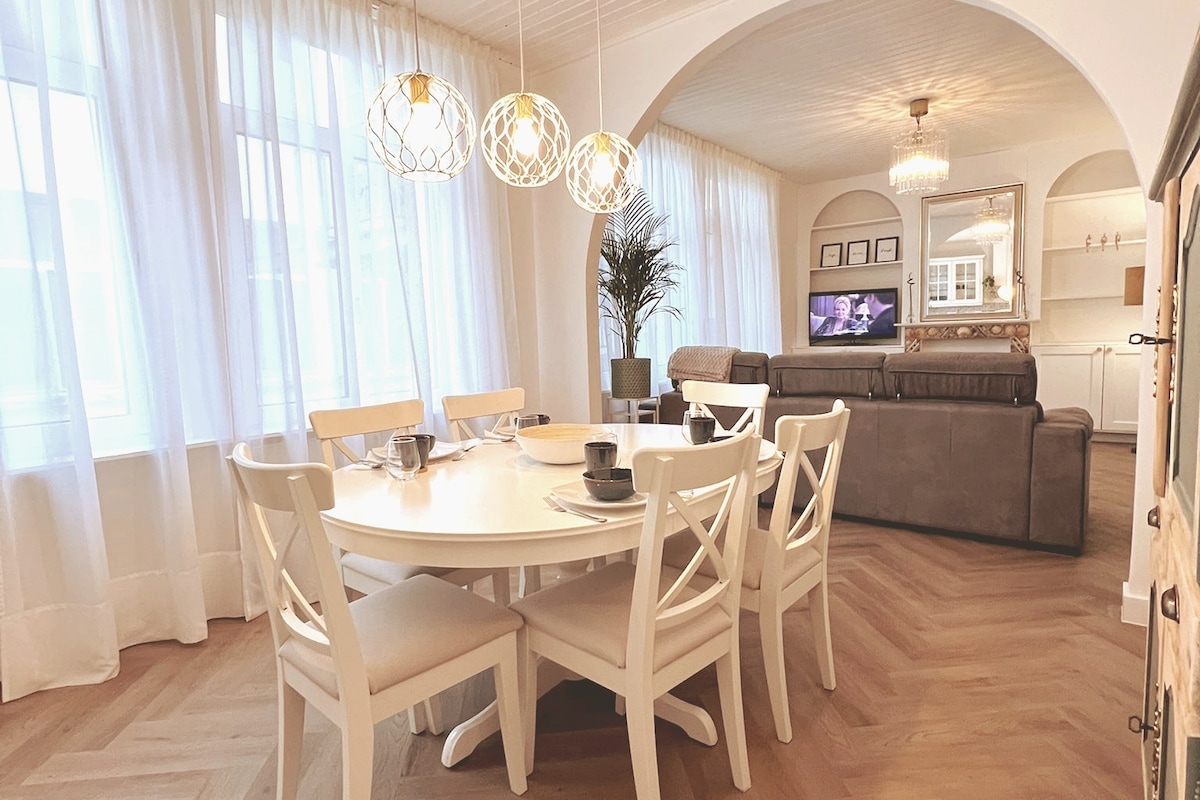 A dining area features a round table surrounded by six white chairs. Above, three pendant lights provide soft illumination. The open space includes a comfortable sofa in the background, with a television visible. Sheer curtains allow natural light to fill the room, enhancing the airy ambiance.