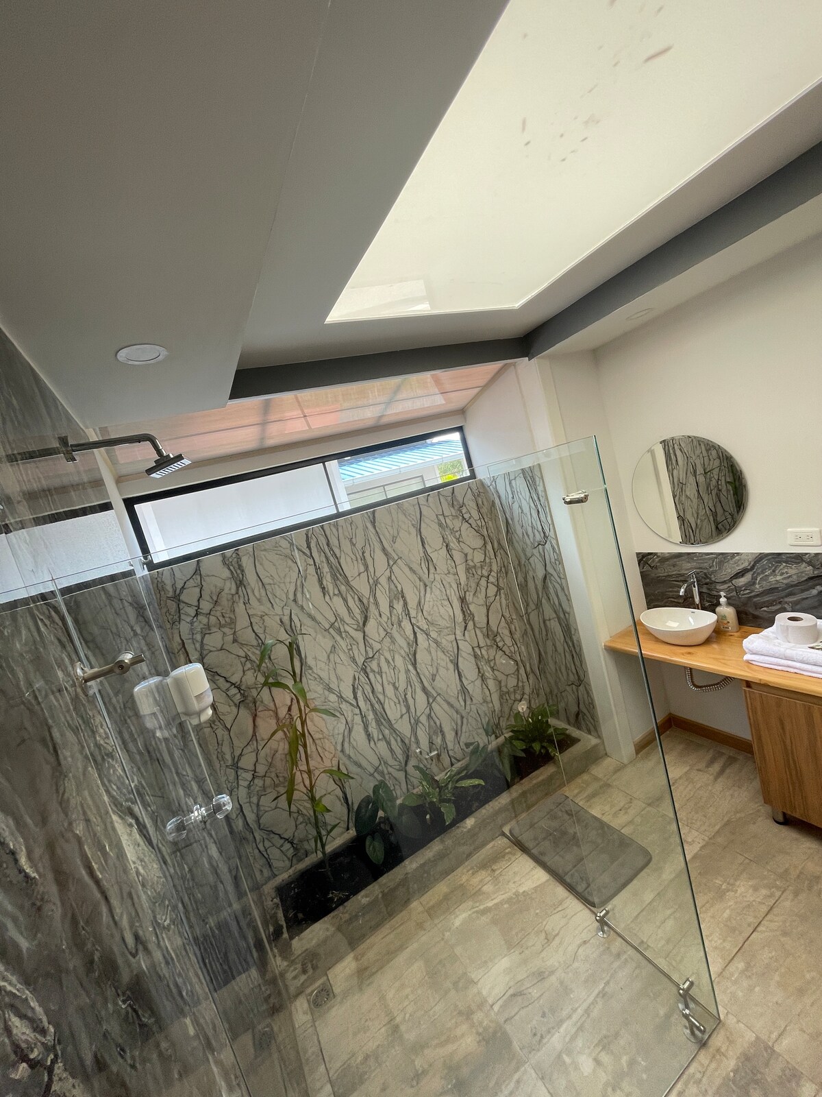 A spacious bathroom features a walk-in shower enclosed by glass panels. Natural light enters through a skylight above, illuminating the patterned stone walls. A wooden vanity with a round mirror reflects a serene environment, complemented by small indoor plants positioned nearby.