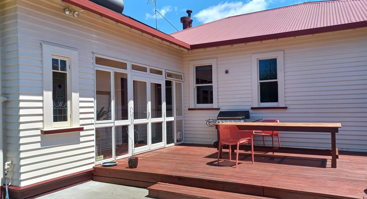 A spacious deck area features a large wooden table accompanied by red chairs, set against a backdrop of the bungalow’s white and red exterior. Large glass doors lead inside, allowing natural light to brighten the space. A barbecue grill is positioned nearby, enhancing outdoor dining options.