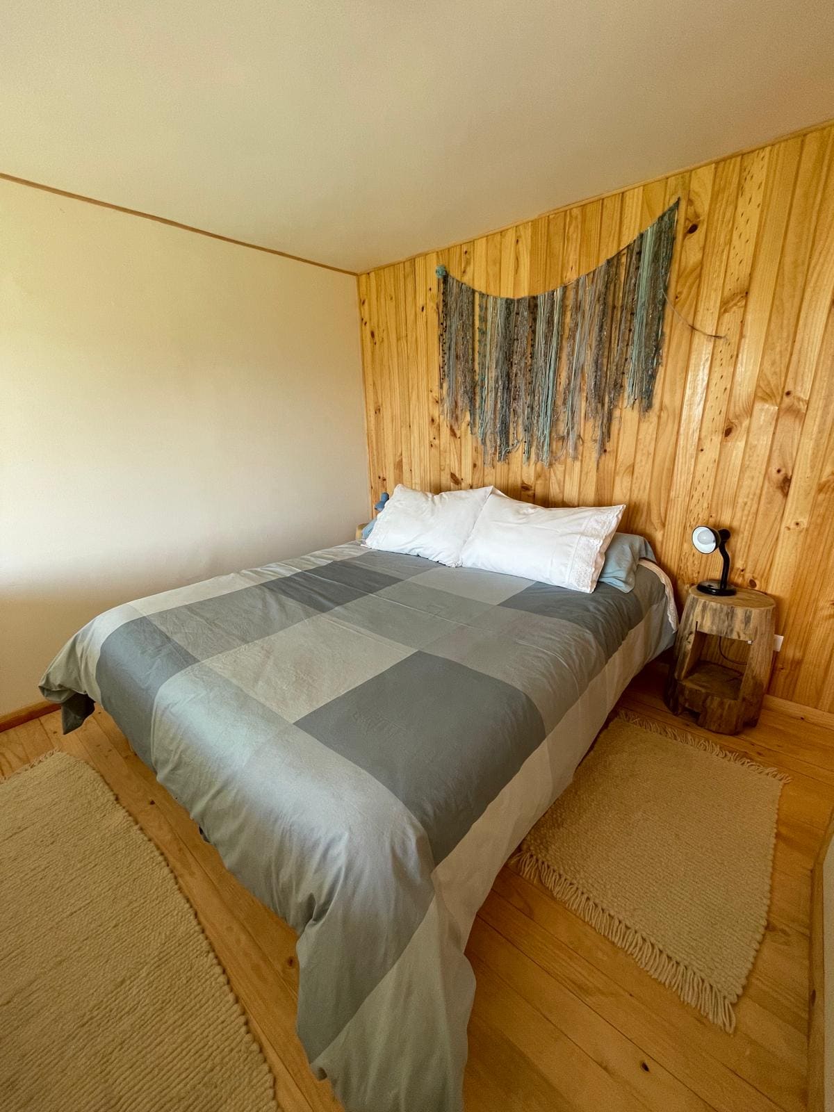 A comfortable sleeping area features a spacious bed with a gray and white patterned coverlet. Natural wood paneling adds warmth to the room, complemented by a decorative wall hanging. A bedside table with a lamp is positioned next to the bed, enhancing the cozy atmosphere.