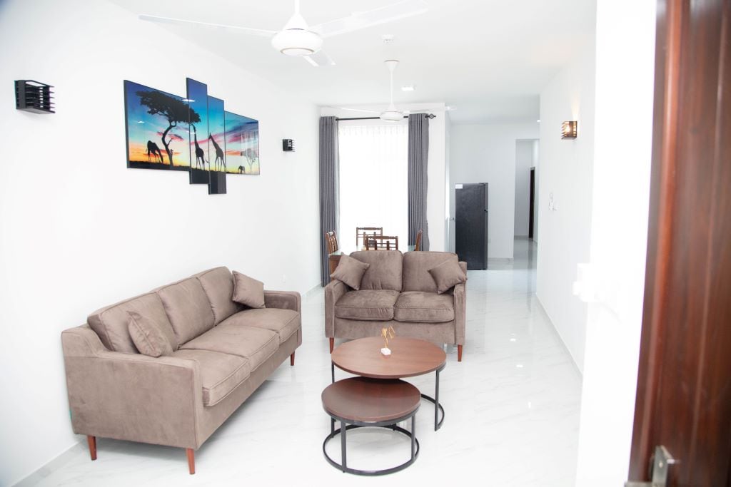 A modern living area showcases a light-hued floor and walls. Two comfortable brown sofas are arranged around a circular coffee table. A contemporary wall art piece depicting a savanna scene adds interest, while a dining area and kitchen are seen in the background.