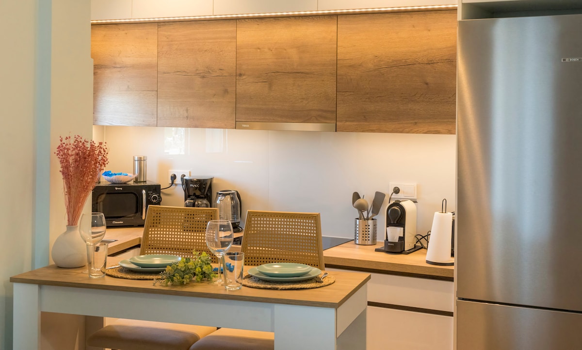 A modern kitchen is showcased with wooden cabinetry and sleek appliances. A cozy dining area is present with a simple table set for two, accompanied by glassware and decorative touches, including a small vase and fresh greenery, creating an inviting space for meals.