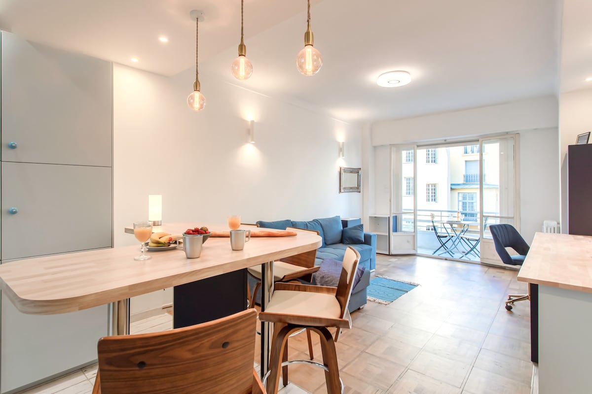 An open-concept living area is featured, with a light wooden kitchen island and bar stools. A comfortable seating area with a blue sofa is visible, alongside a small balcony that offers natural light. A table with drinks and fruit adds to the inviting space.