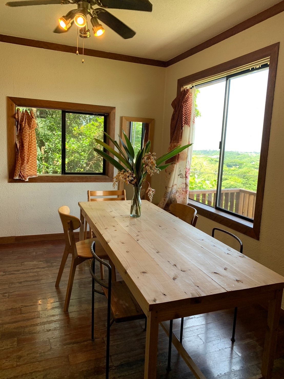 A spacious dining area is highlighted by a wooden table surrounded by chairs. Large windows offer views of the lush greenery outside, allowing natural light to fill the room. A vase with fresh plants adds a decorative touch to the table.