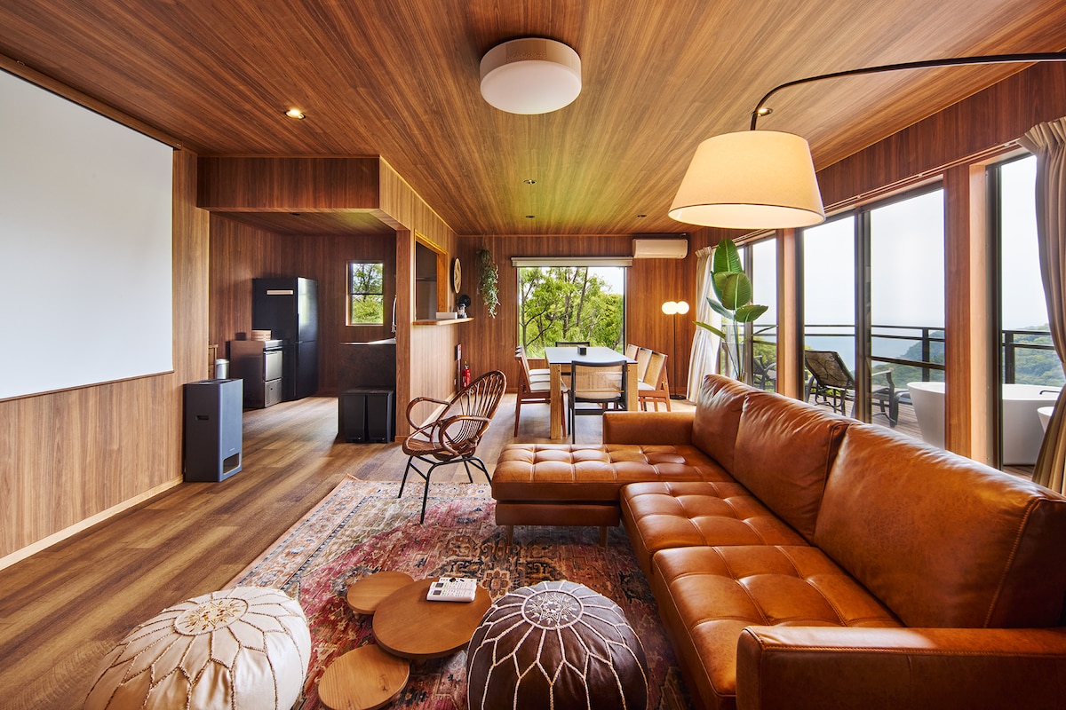 A spacious living area features wooden accents and large windows, allowing natural light to fill the space. A comfortable leather sofa is complemented by a wooden coffee table and ornamental cushions. A dining area and modern kitchen appliances are visible in the background.