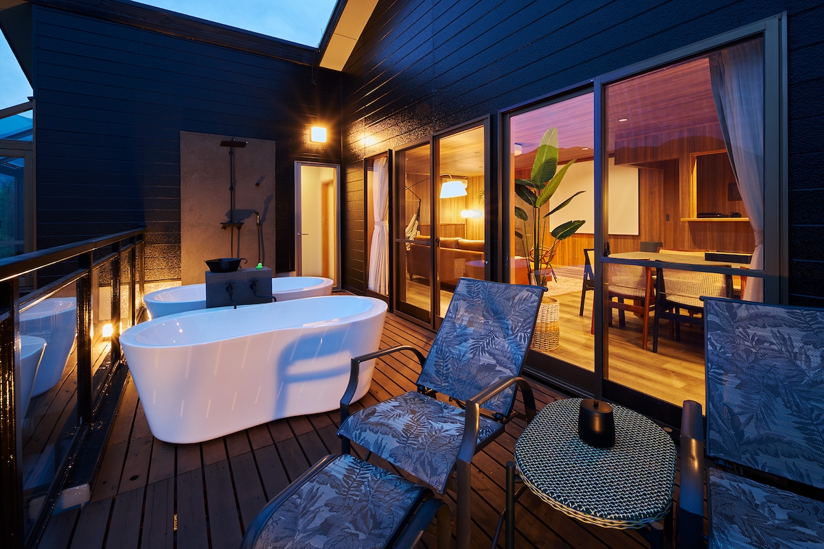 An outdoor deck area is showcased, featuring a stylish freestanding bathtub alongside two lounging chairs. Soft lighting enhances the surroundings, which include a table and chairs in the background, creating an inviting space for relaxation.