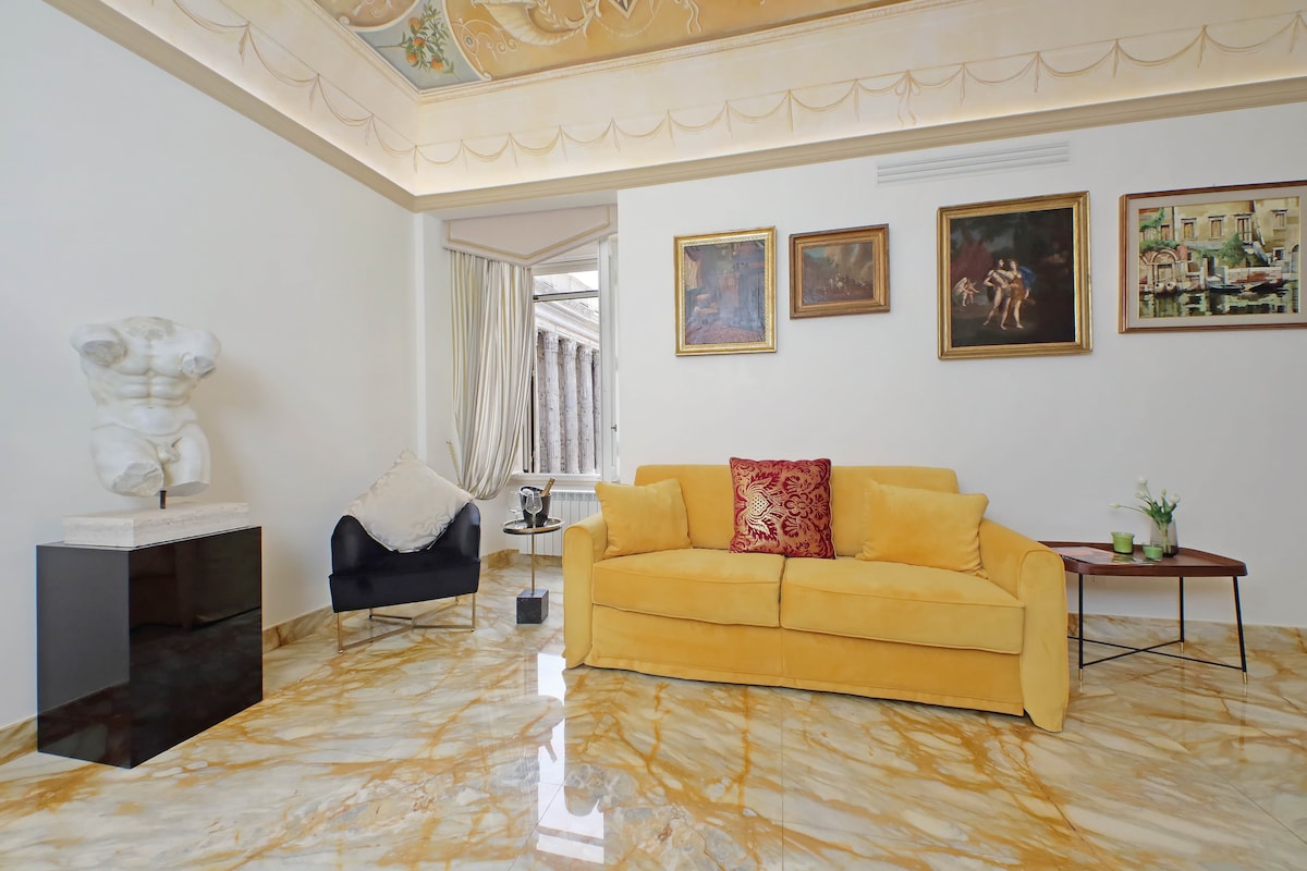 A bright living area features a plush yellow couch paired with a dark accent chair. Artistic wall decorations, including framed paintings, enhance the space. A marble floor reflects the light, while a classical statue adds a touch of elegance near the seating.