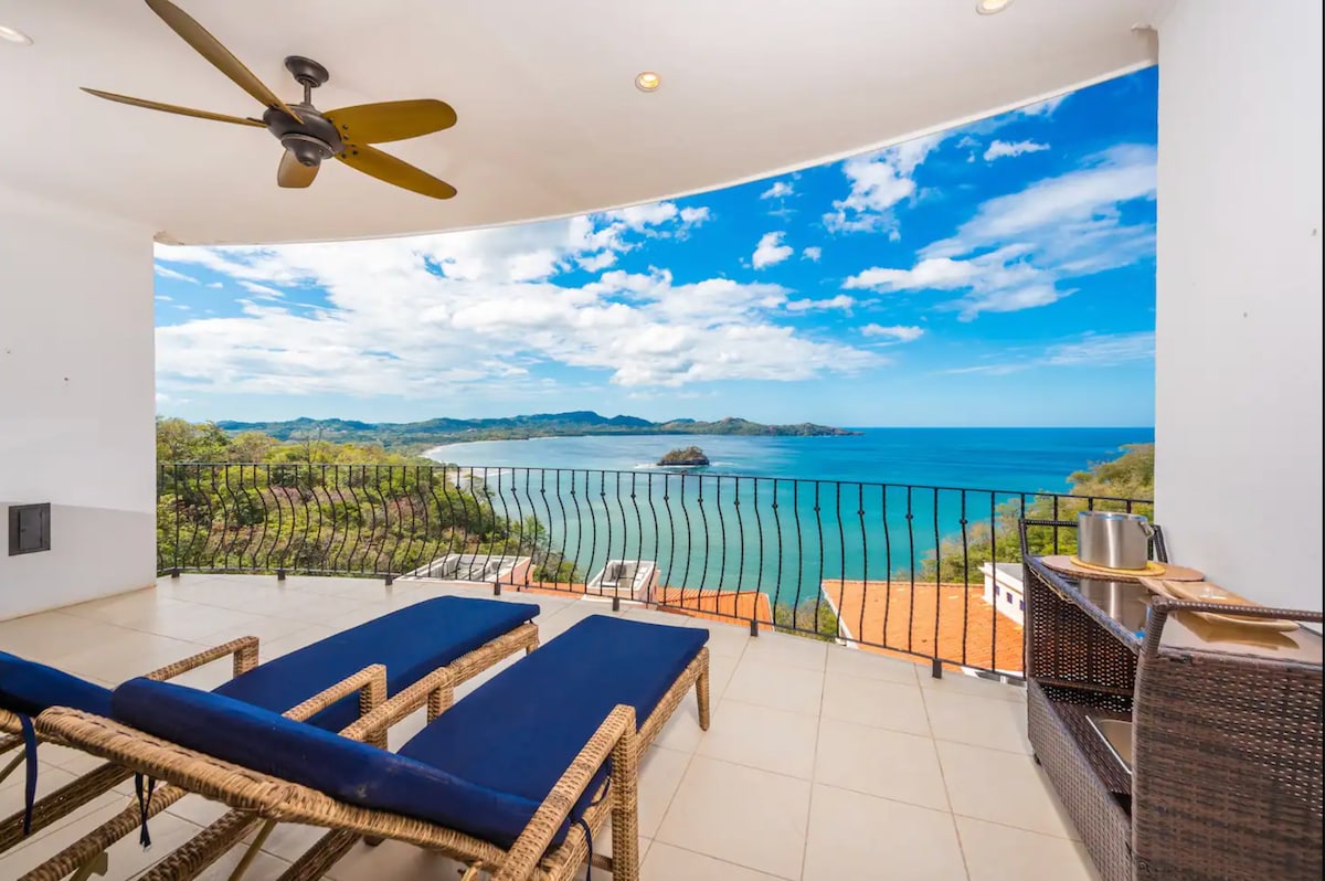 A spacious private deck overlooks the Pacific Ocean, featuring two lounge chairs and a small table. The scene captures vibrant blue waters and distant hills under a partly cloudy sky, creating an inviting space for relaxation.