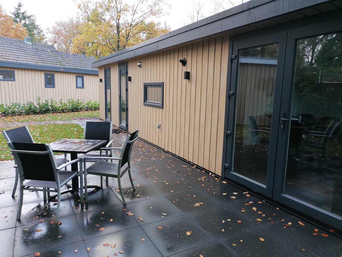 A terrace area is shown, featuring a dining table with four matching chairs. The structured wooden walls of the chalet can be seen beside a well-maintained garden space, with fallen leaves scattered across the paved flooring.