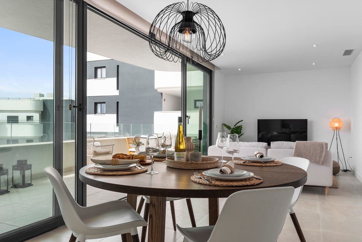A dining area is presented, featuring a round wooden table set with plates and glassware. Large glass doors provide natural light and access to a balcony. A cozy seating area with a light-colored sofa and a modern lamp is visible in the background.