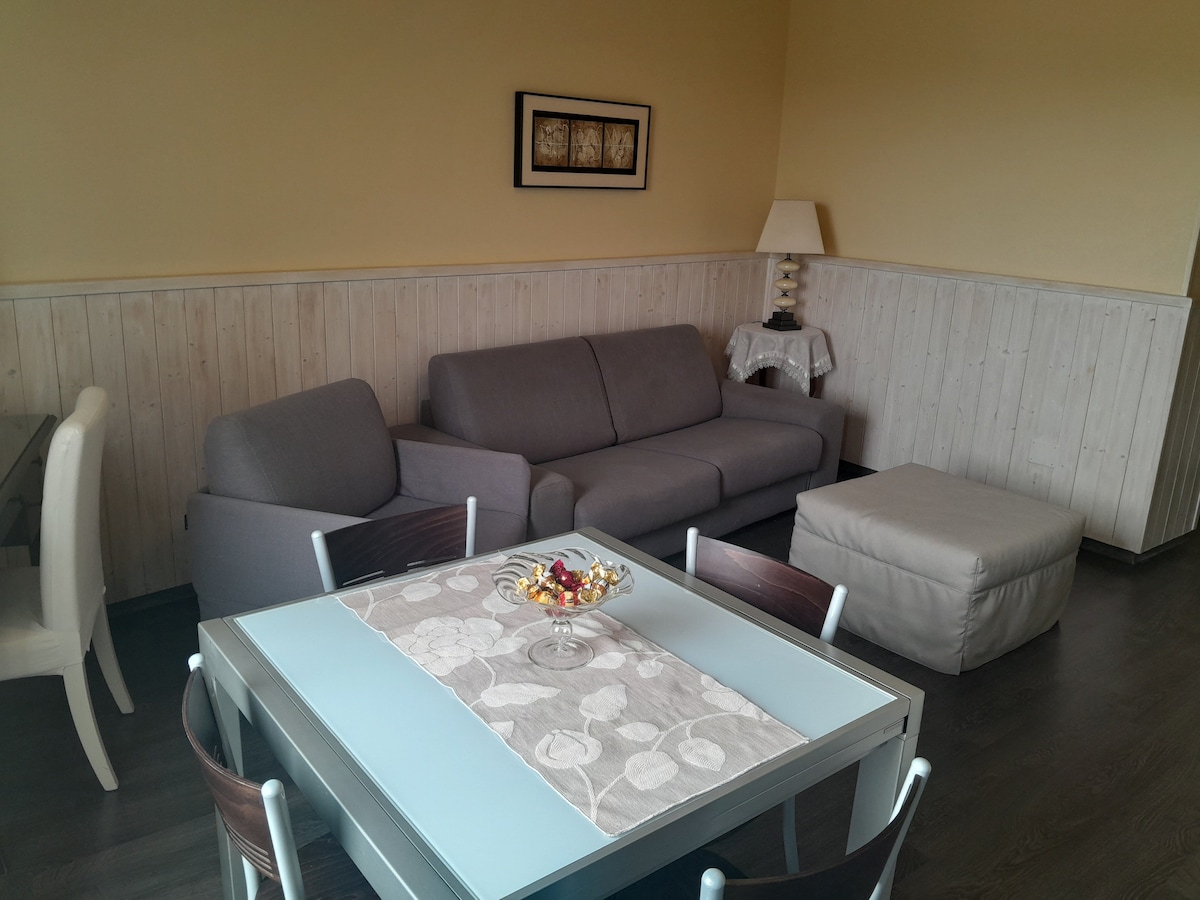 A living area features a gray sectional sofa and a light-colored ottoman. A wooden dining table is set with a decorative centerpiece, surrounded by four dark chairs. The walls are partially clad in light wood, and natural light fills the space.