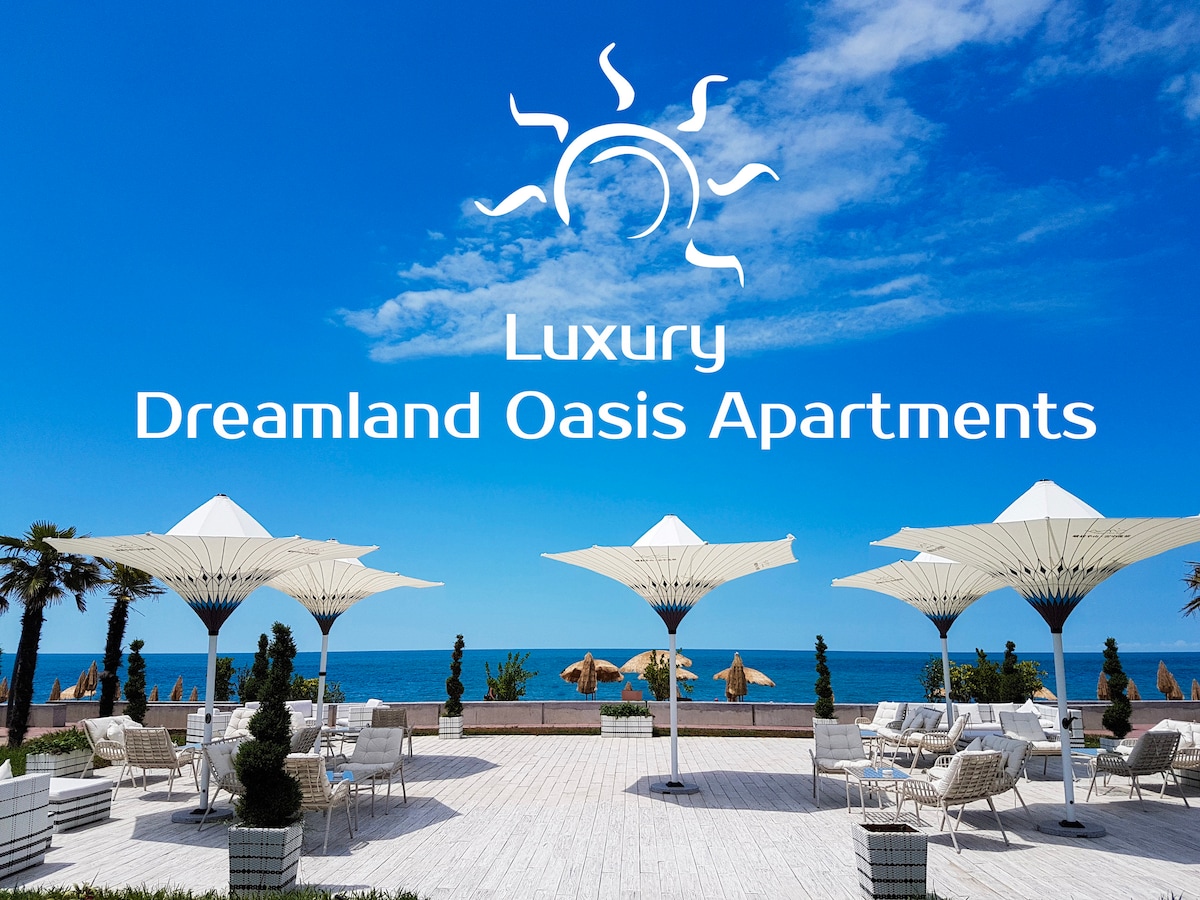 An outdoor lounge area features white patio furniture under large umbrellas, providing shade. In the background, the ocean meets a clear blue sky, while palm trees frame the space, adding to the relaxed coastal ambiance.