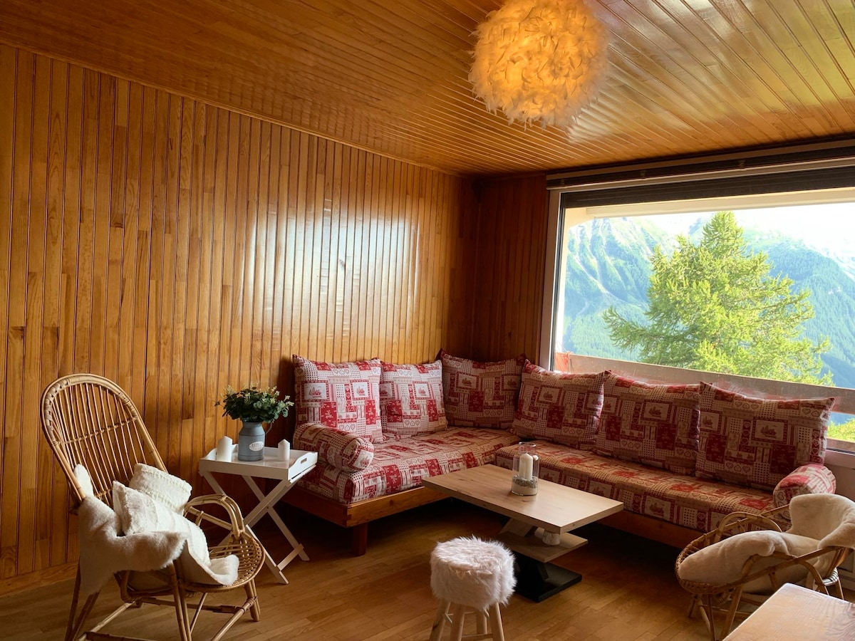 A cozy living area features wooden wall paneling and a large, comfortable couch with patterned cushions. A small coffee table is placed in the center, accompanied by a side table. A sizable window offers a view of the surrounding mountains, complemented by natural light.