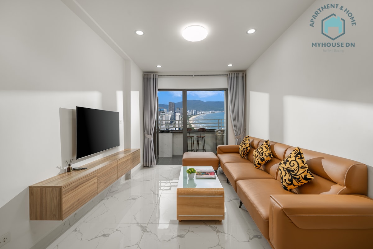 A modern living area features a spacious segmented orange couch with decorative cushions, facing a wall-mounted television. A wooden media console is positioned below the TV. Natural light floods the space through large windows, offering views of the cityscape and distant mountains.