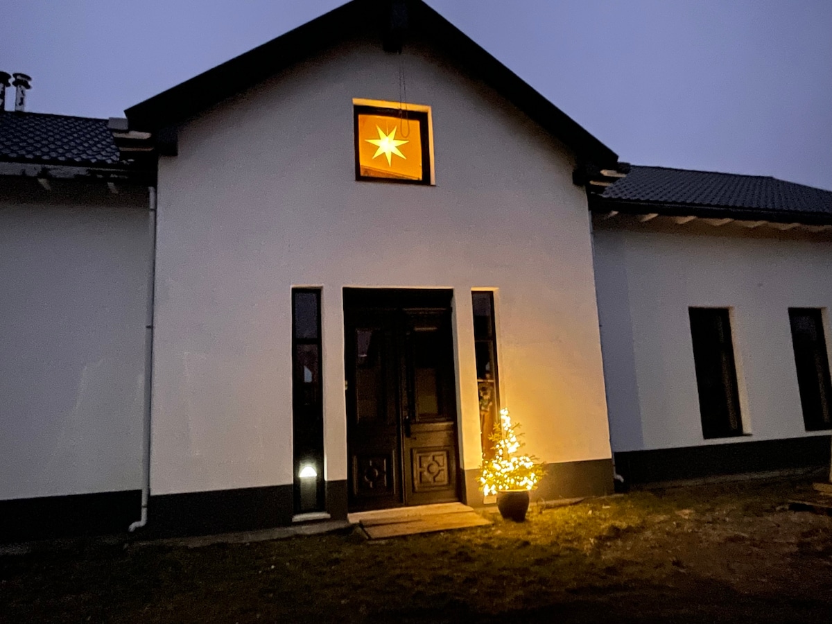 The exterior of the accommodation features a bright star decoration above the entrance, with a small tree lit up beside the door. The structure is white with dark accents, and large windows allow for ample natural light.