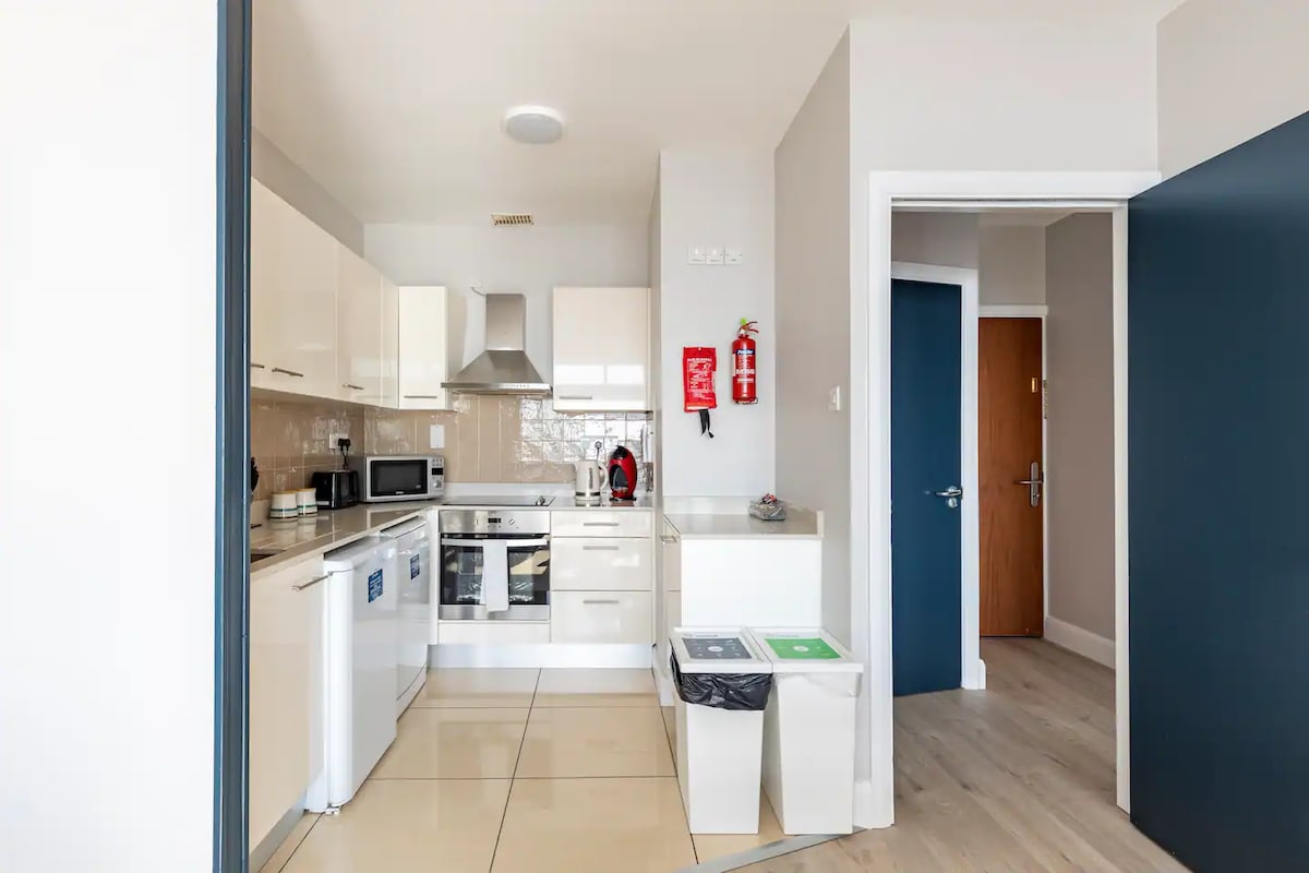 A modern kitchen is equipped with sleek white cabinetry and stainless steel appliances, including a microwave and dishwasher. Bright tiles reflect natural light, enhancing the space. A door on the right leads to an adjoining room, and a recycling bin is positioned near the entrance.