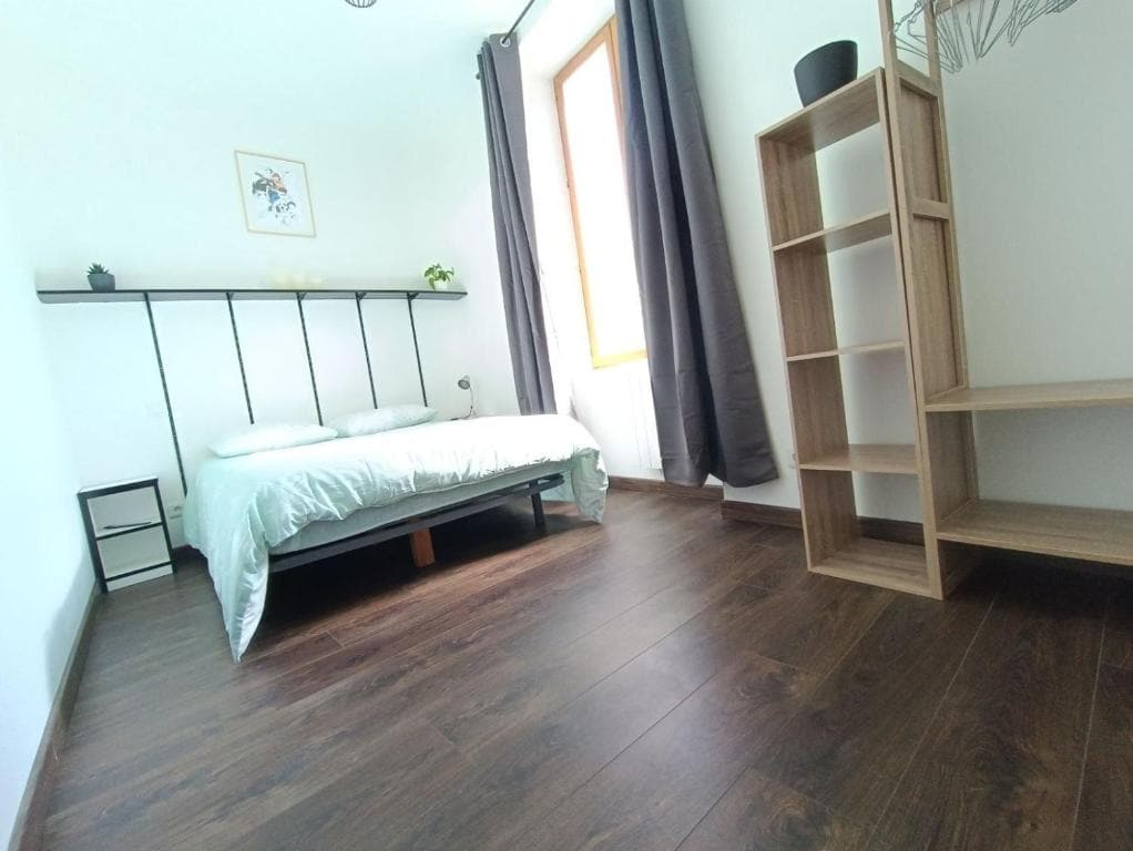 A bright bedroom features a spacious bed with a light-colored comforter positioned against a wall. A simple shelf and a modern bookshelf stand nearby, while a window with dark curtains allows natural light to enter the room, enhancing the wooden flooring.