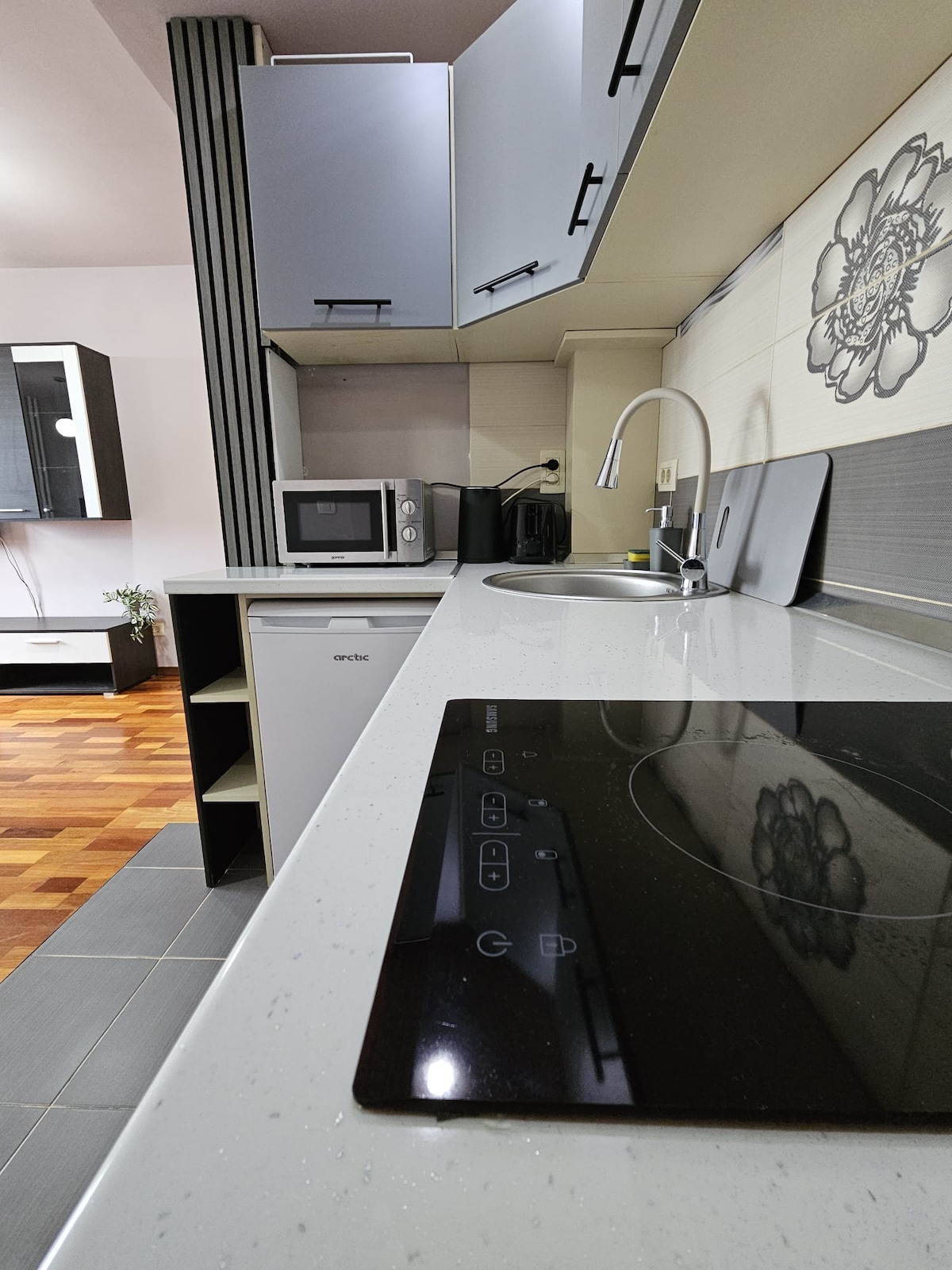 A modern kitchen is presented, featuring a sleek countertop and an integrated stove. The cabinetry above offers storage, while a microwave and sink are visible. The flooring includes varied textures, contributing to an organized and functional cooking space.