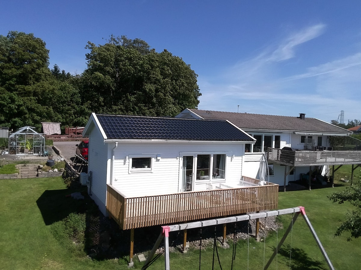 A newly built guesthouse is situated on an expansive lawn, featuring solar panels on the roof. Large windows allow natural light to fill the interior. A wooden deck wraps around the front, providing outdoor space, while a playground can be seen in the background.