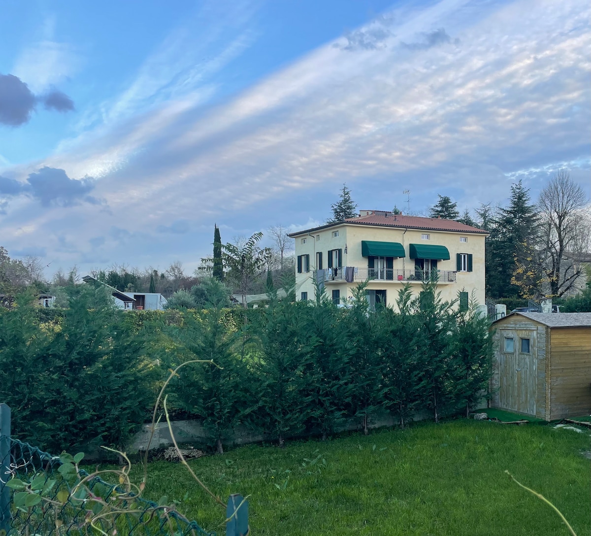 A two-story building is surrounded by neatly trimmed hedges and greenery. The exterior features a light-colored facade with green awnings over the windows. A clear sky with streaks of clouds is visible above, contributing to the peaceful outdoor setting.