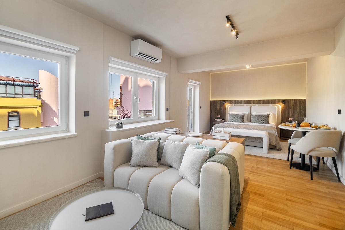 A spacious studio is showcased with large windows allowing natural light. A comfortable seating area with a light sofa and small round table complements the space. The stylish double bed with soft linens is visible in the background, creating a relaxed ambiance.