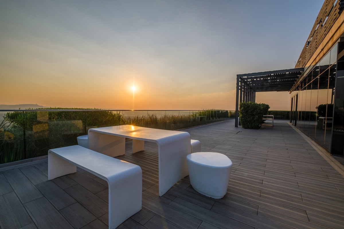 A spacious rooftop terrace is presented with modern white furniture, including a rectangular table and round stools. Lush greenery borders the area, while a stunning sunset casts a warm glow over the horizon, creating a serene outdoor setting.