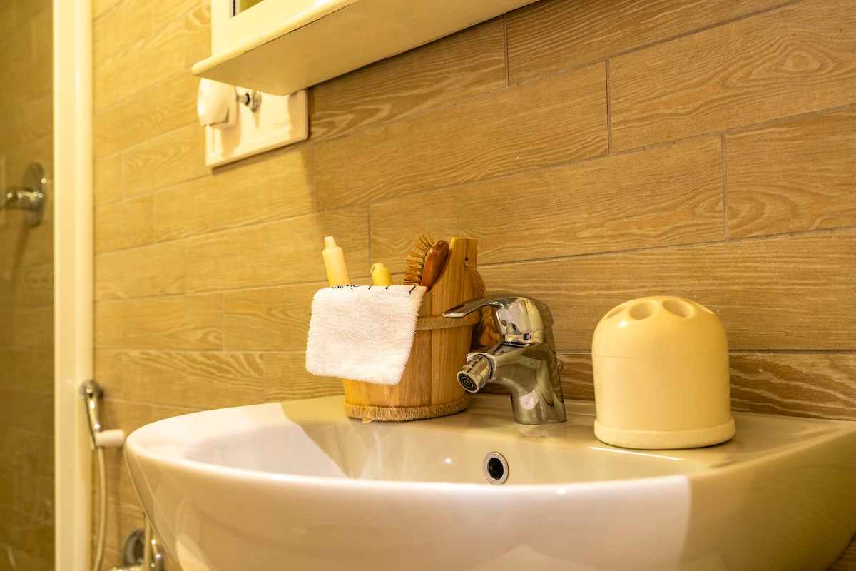 A modern bathroom sink is paired with a wooden storage basket, containing toiletries such as lotions and brushes. The sink features a contemporary faucet, while a soap dispenser is present nearby. The warm-toned wood paneling provides a cozy backdrop.