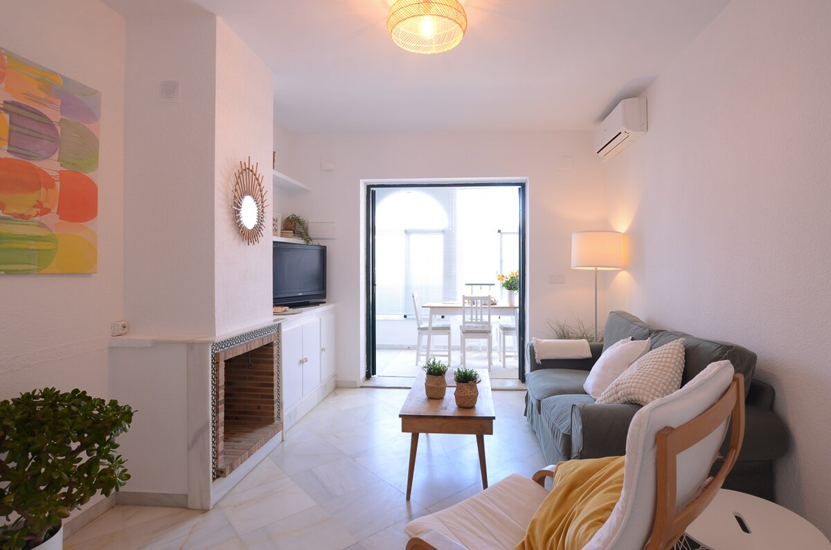 A comfortable living area features a green sofa, accent chair, and a coffee table with plants. Natural light floods the space through large glass doors that lead to a terrace with outdoor seating, creating a bright and inviting atmosphere.