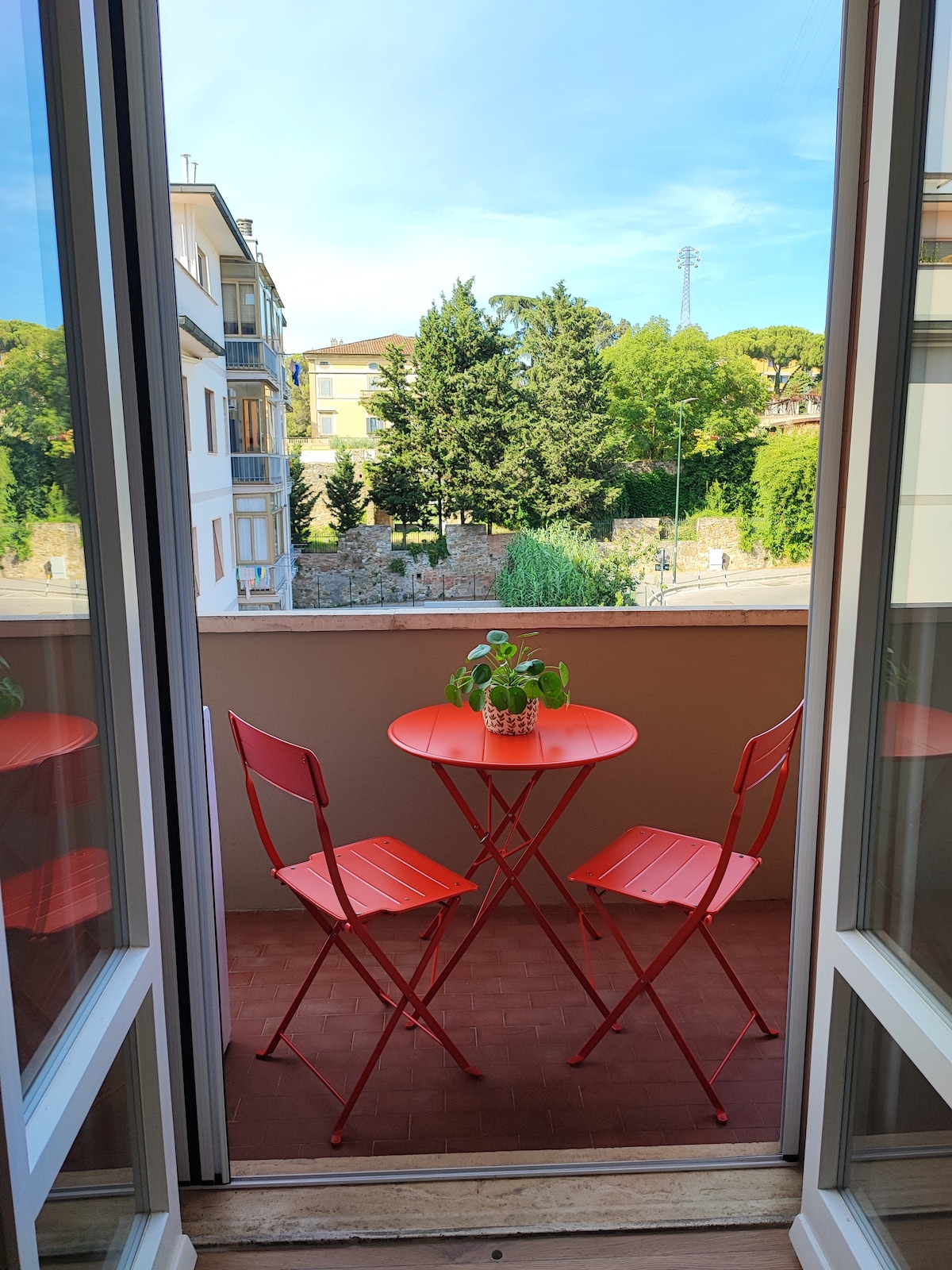 A small balcony features a round orange table with two matching chairs, surrounded by greenery. The view offers glimpses of trees and residential buildings beyond. Natural light fills the space through the open double doors, creating a relaxed outdoor atmosphere.