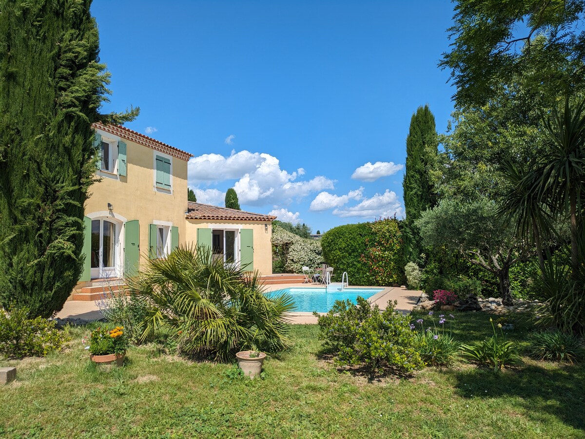 A two-story yellow villa sits surrounded by greenery, featuring a private swimming pool in the foreground. Lush plants border the pool area, while the building showcases green shutters and terracotta roofing. A clear blue sky complements the tranquil outdoor setting.