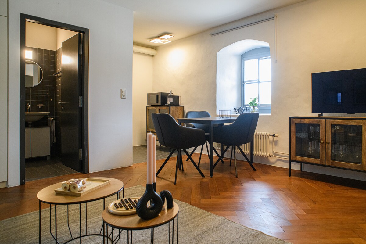 A spacious living area features two round tables accompanied by black chairs. In the background, a kitchenette with modern appliances is visible. A cozy lounge area includes two small tables, one holding candles and a decorative item. Natural light enters through a window, enhancing the warm atmosphere.