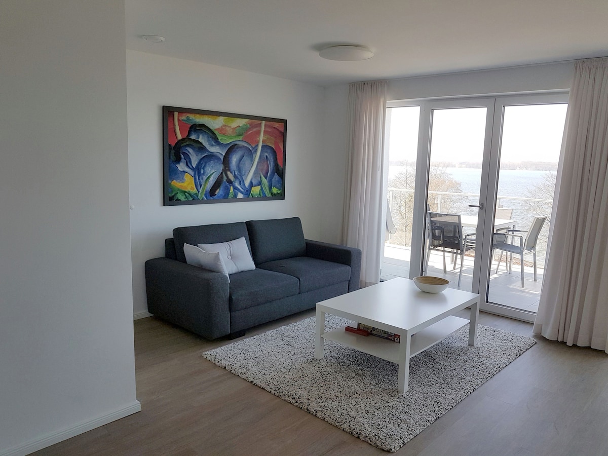 A living area is depicted with a gray sofa positioned against a wall, complemented by a colorful artwork above it. A white coffee table rests on a textured rug, while large glass doors lead to a balcony overlooking the lake. Natural light fills the space.