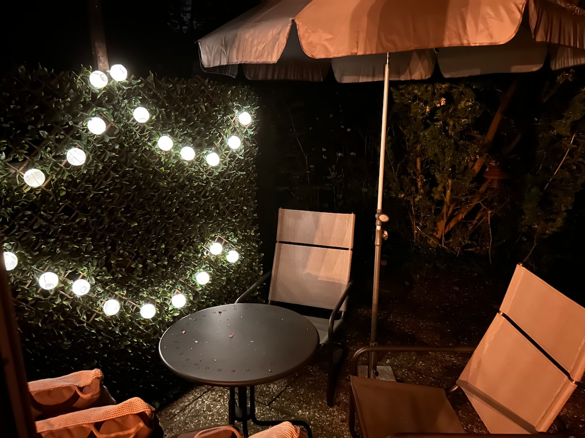 A peaceful garden space is illuminated by soft string lights. Two light-colored lounge chairs and a round table are positioned beneath a large umbrella, creating a cozy atmosphere for relaxation. Lush greenery forms a backdrop, enhancing the inviting setting.